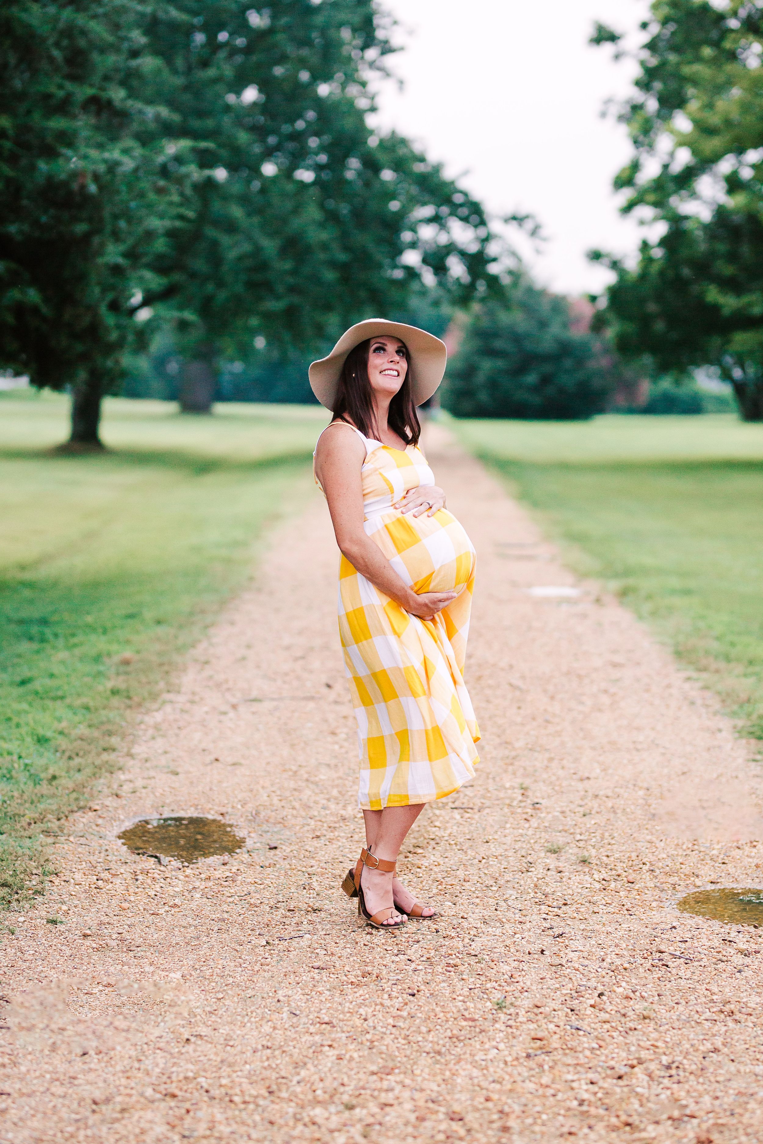 Smiling pregnant brunette in yellow plaid dress and sun hat on a gravel garden path.