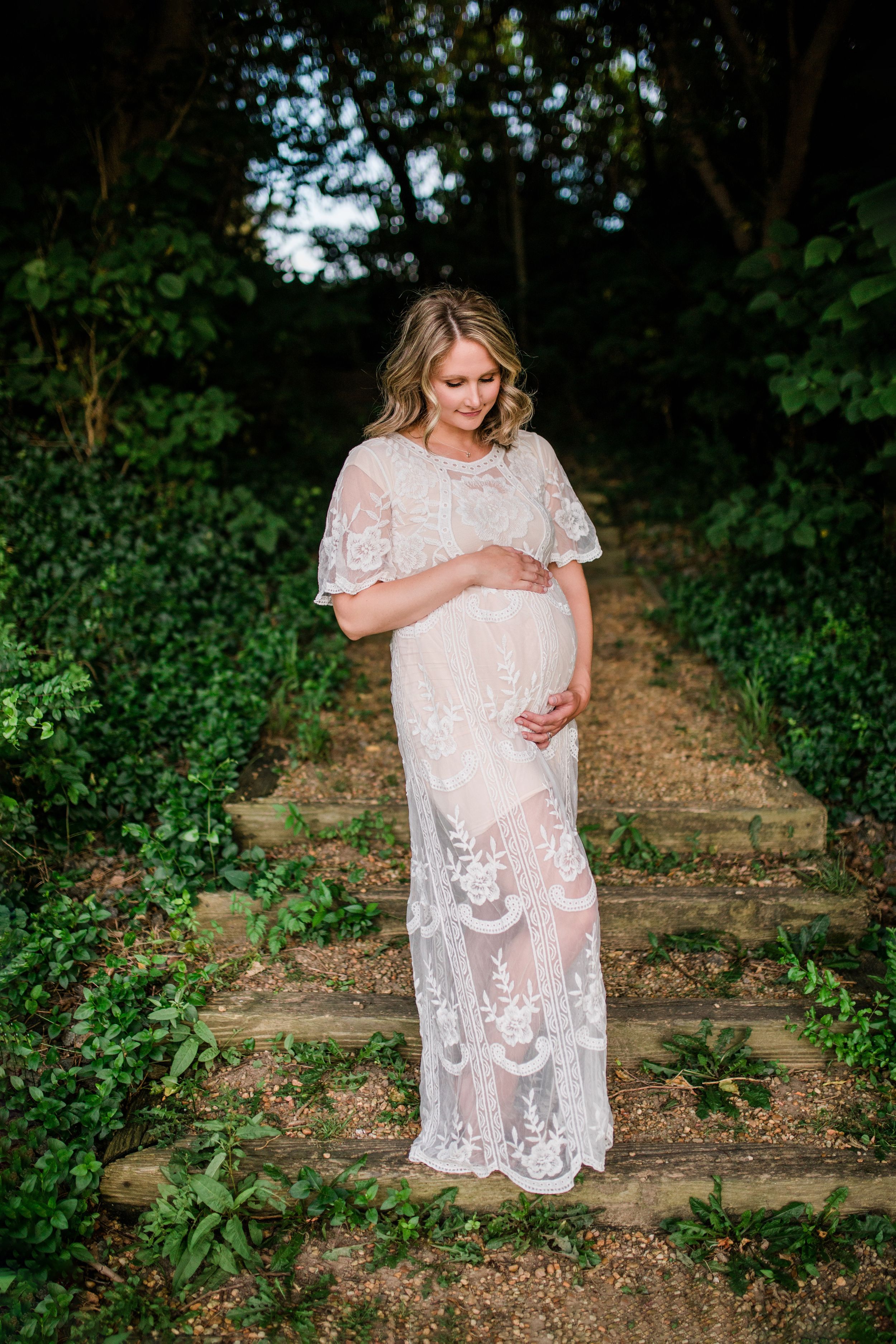 Pregnant woman in sheer white lace dress standing on garden steps surrounded by lush foliage.