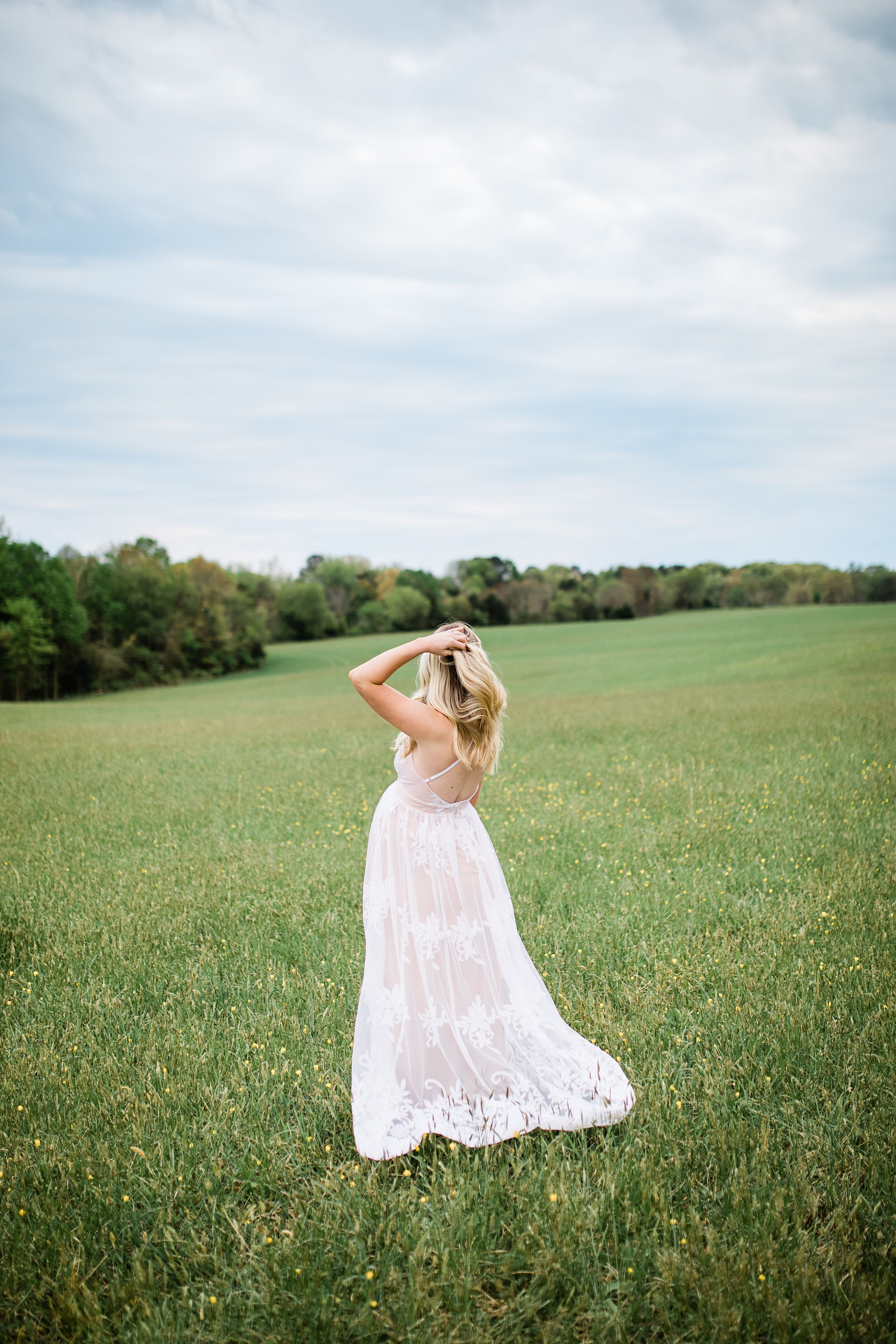 Pregnant woman in flowing white lace dress standing in open green field under cloudy sky.