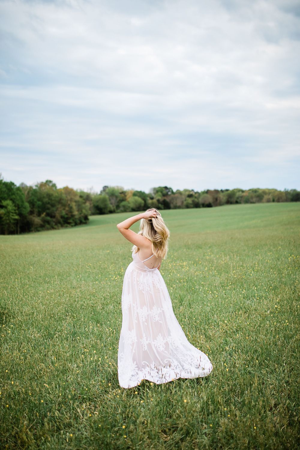Pregnant woman in flowing white lace dress standing in open green field under cloudy sky.