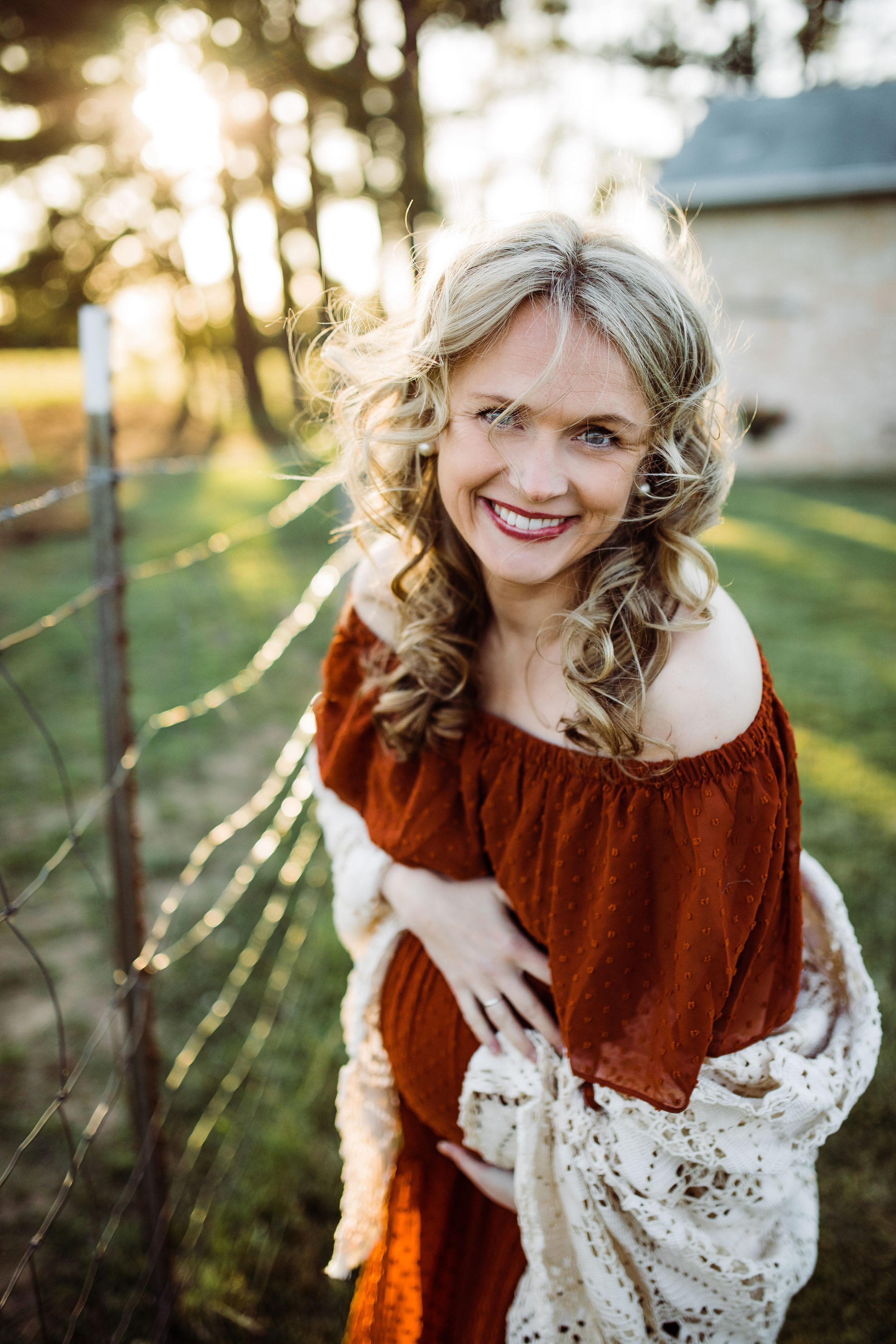 Smiling blonde pregnant woman in rust off-shoulder dress posing near barbed wire fence at sunset.