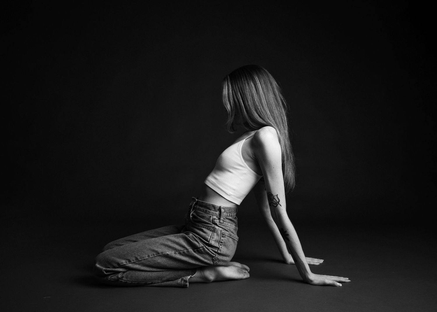 Minimalist black and white portrait of figure kneeling in casual attire against dark background.
