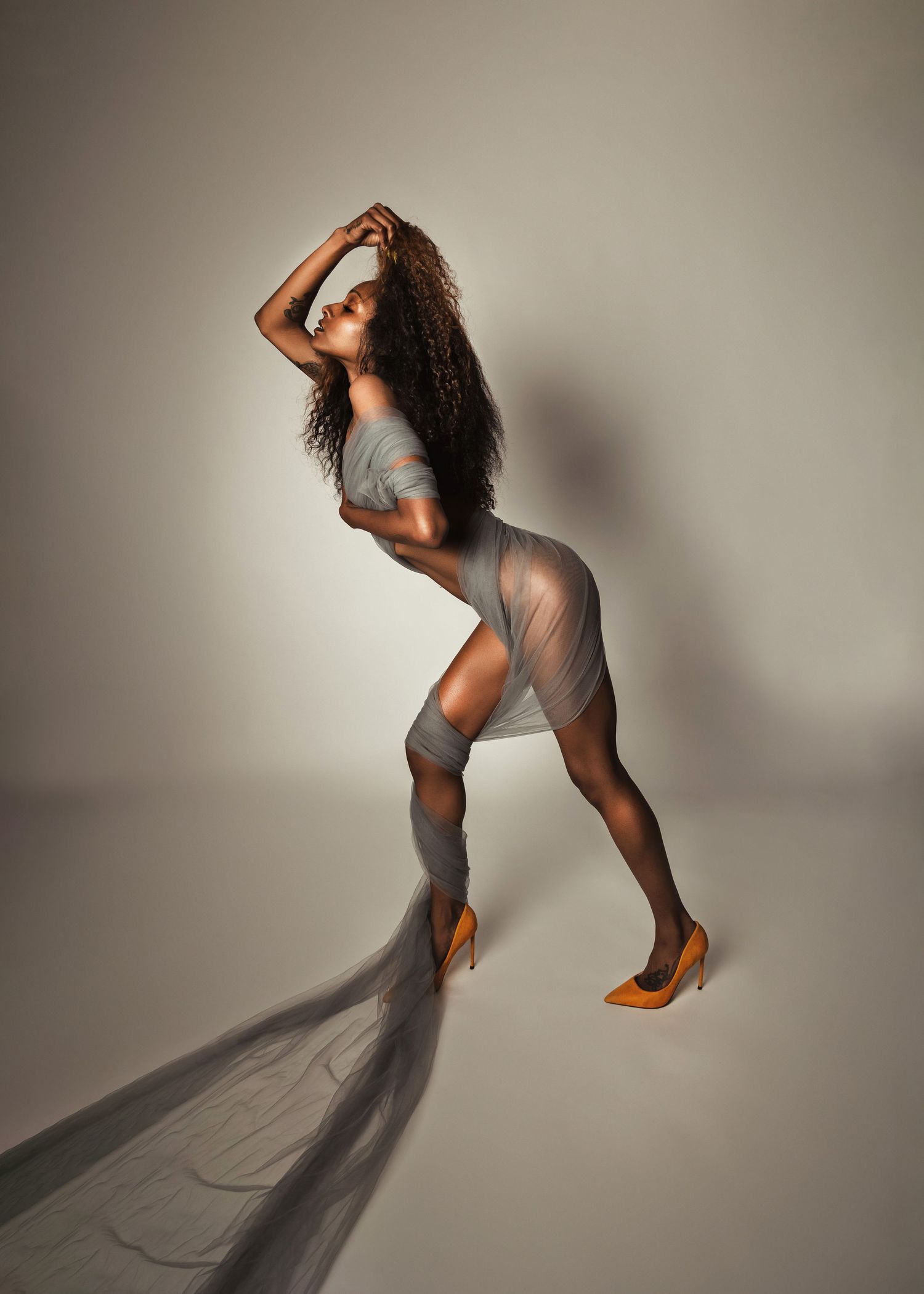 Figure in sheer grey dress striking dramatic pose against neutral studio background.