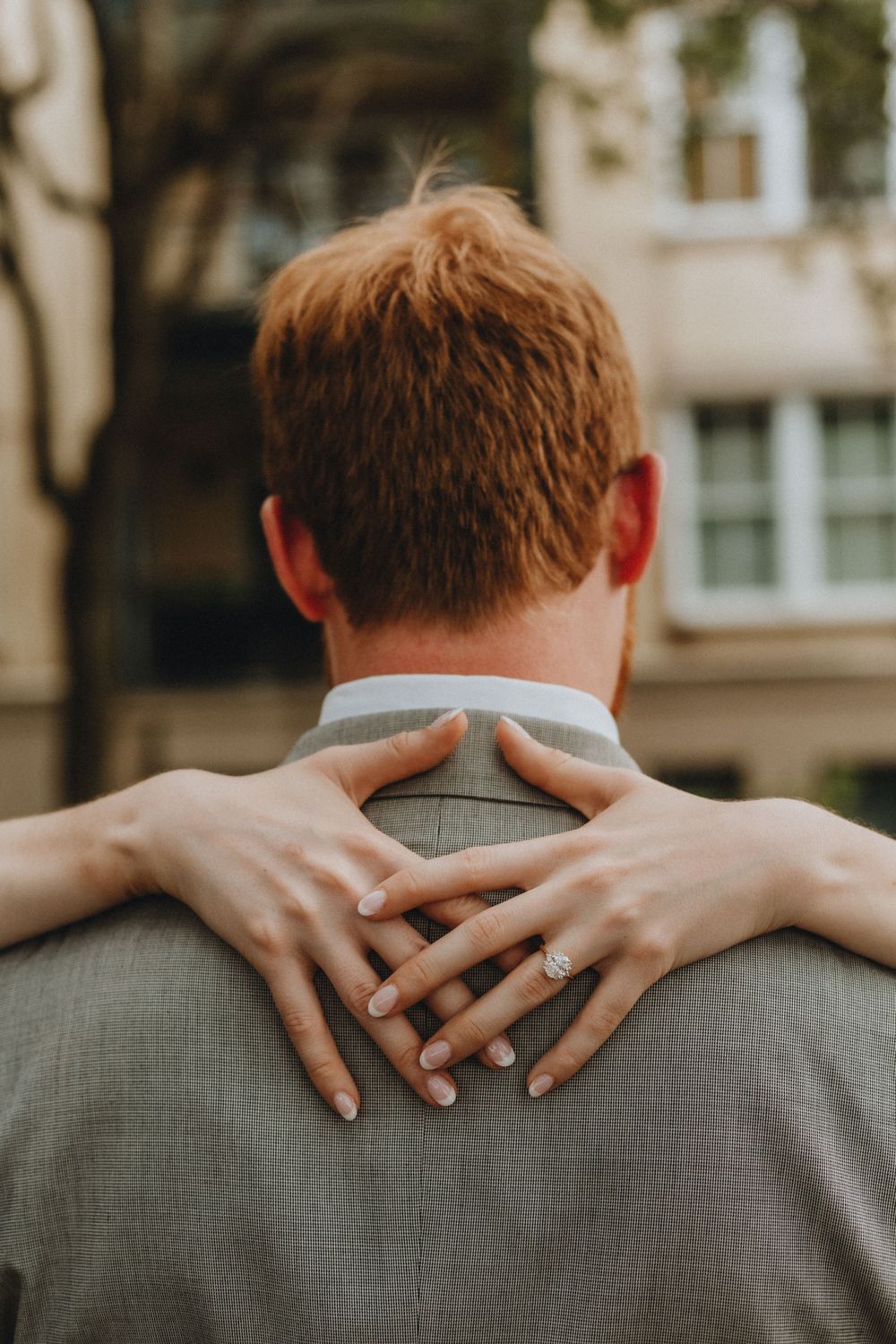 The back of a man's head with a woman's hands on his neck showing off an engagement ring