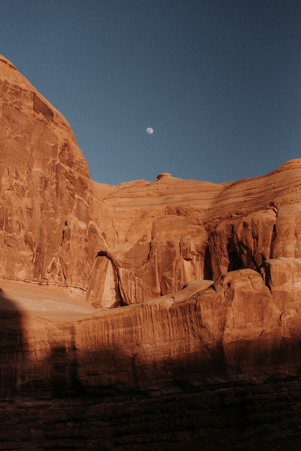 Moon rises over massive red sandstone cliffs in a desert landscape during sunset.