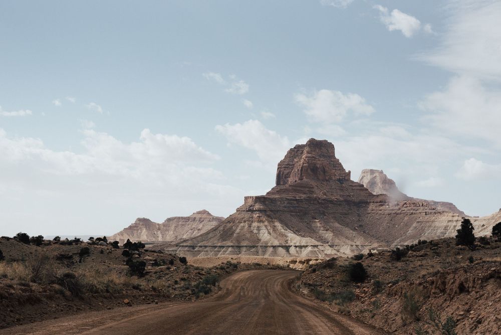Desert road leads to a prominent red rock formation under cloudy skies.
