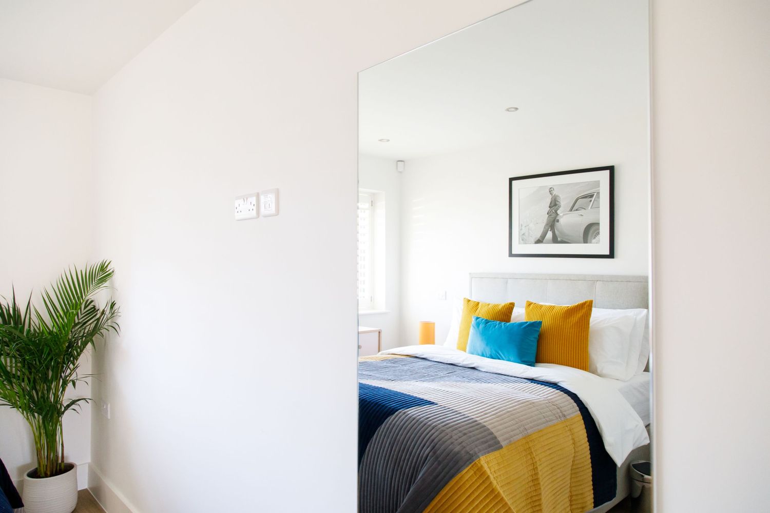 Bright modern bedroom with yellow and gray bedding and a large mirror reflecting natural light.