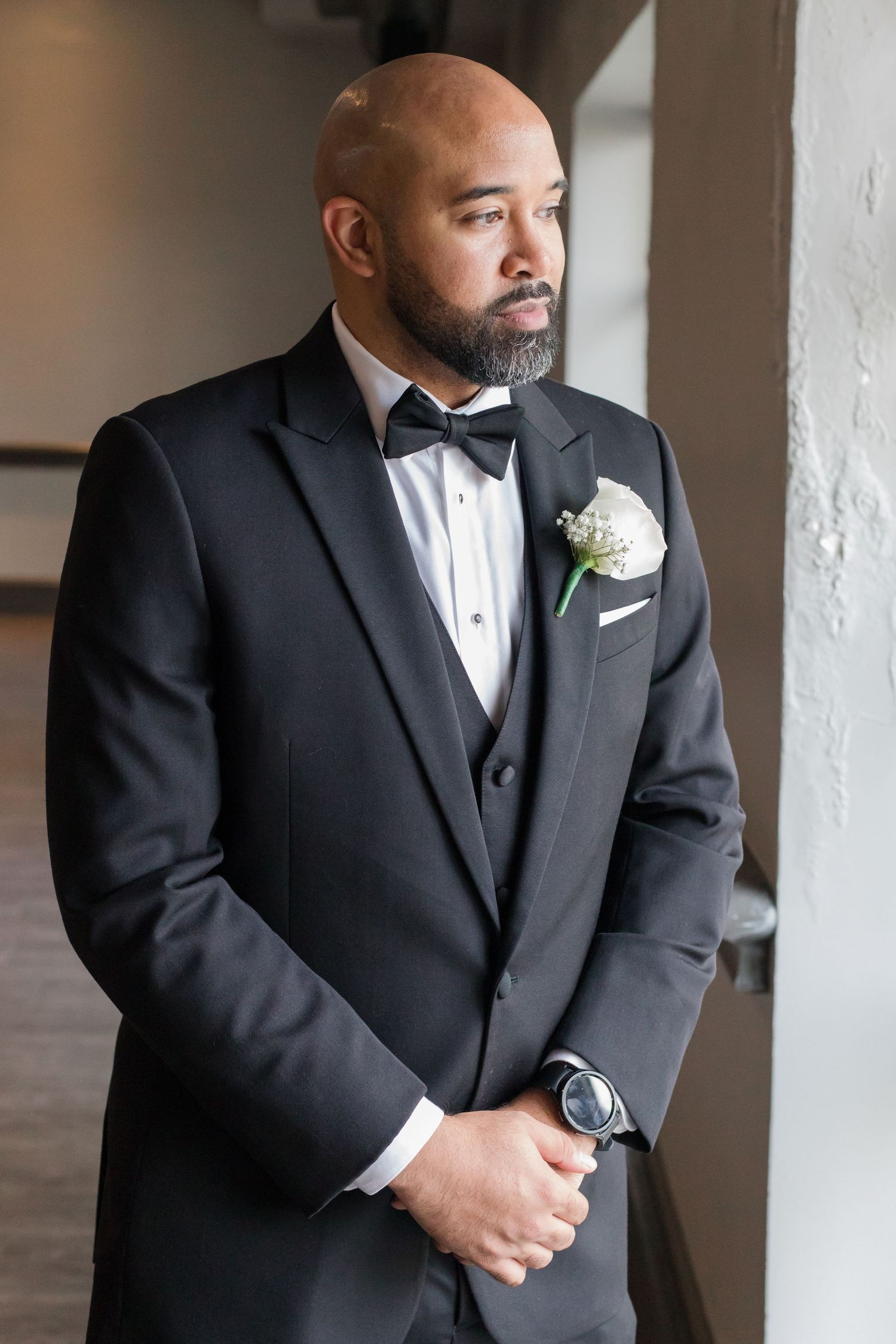 Groom in classic black tuxedo with white boutonniere poses near window.