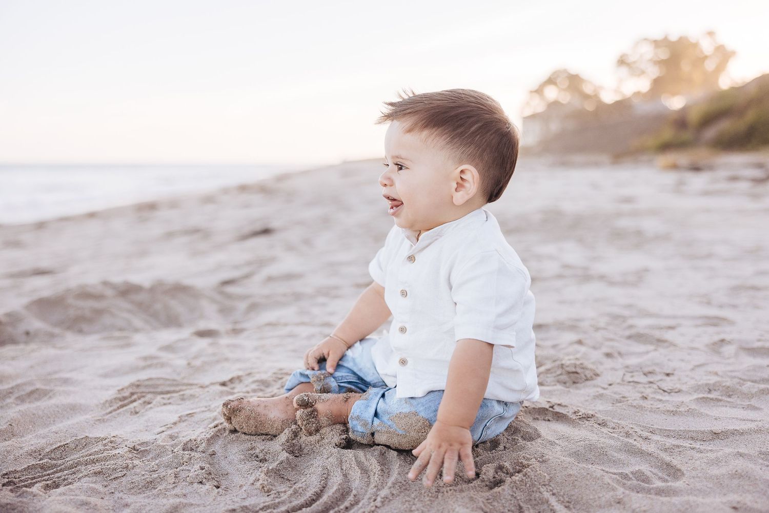 Small child sits on sandy beach wearing white shirt and jeans during sunset.