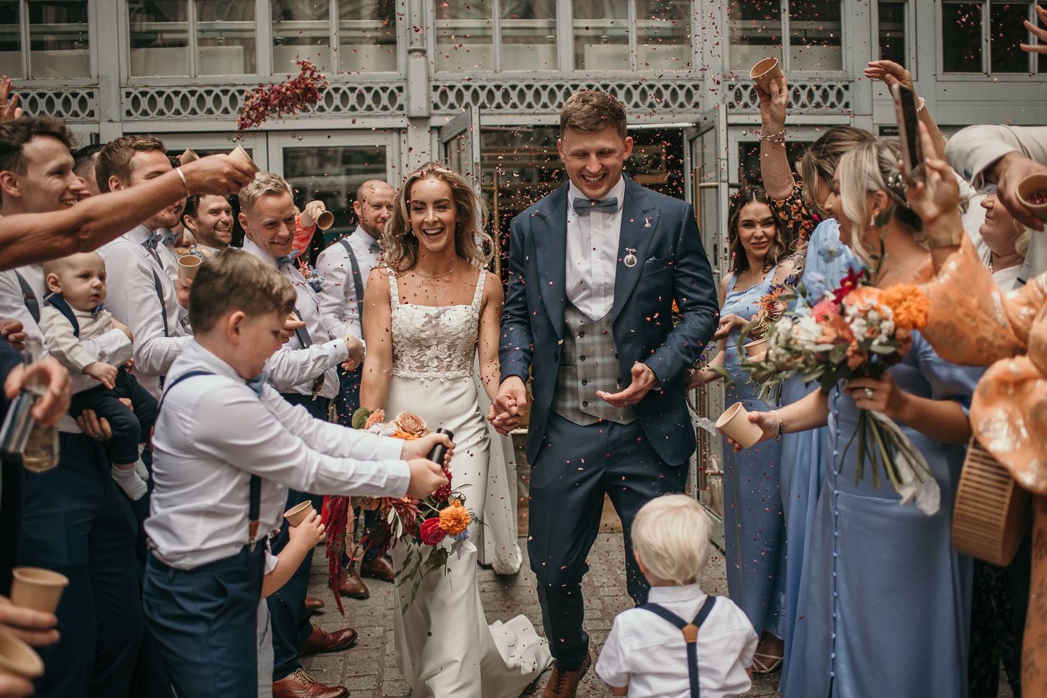 Wedding guests shower the newlyweds with confetti outside a white latticed building.