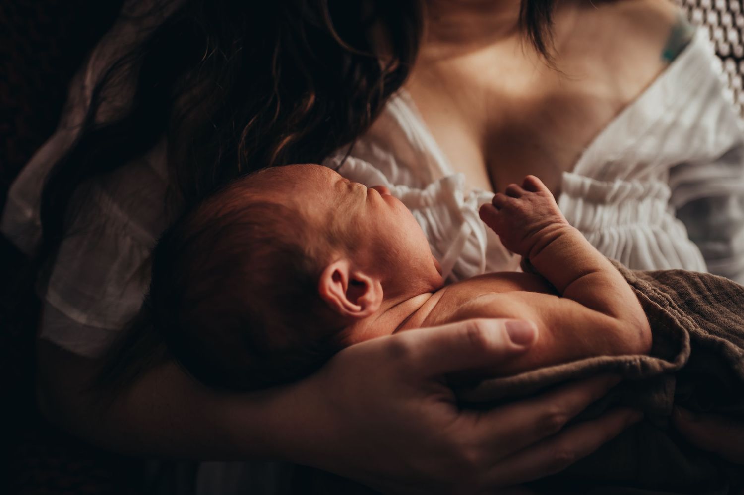mama sits in a peacock chair by a window and prepares to nurse her newborn son at lifestyle newborn session