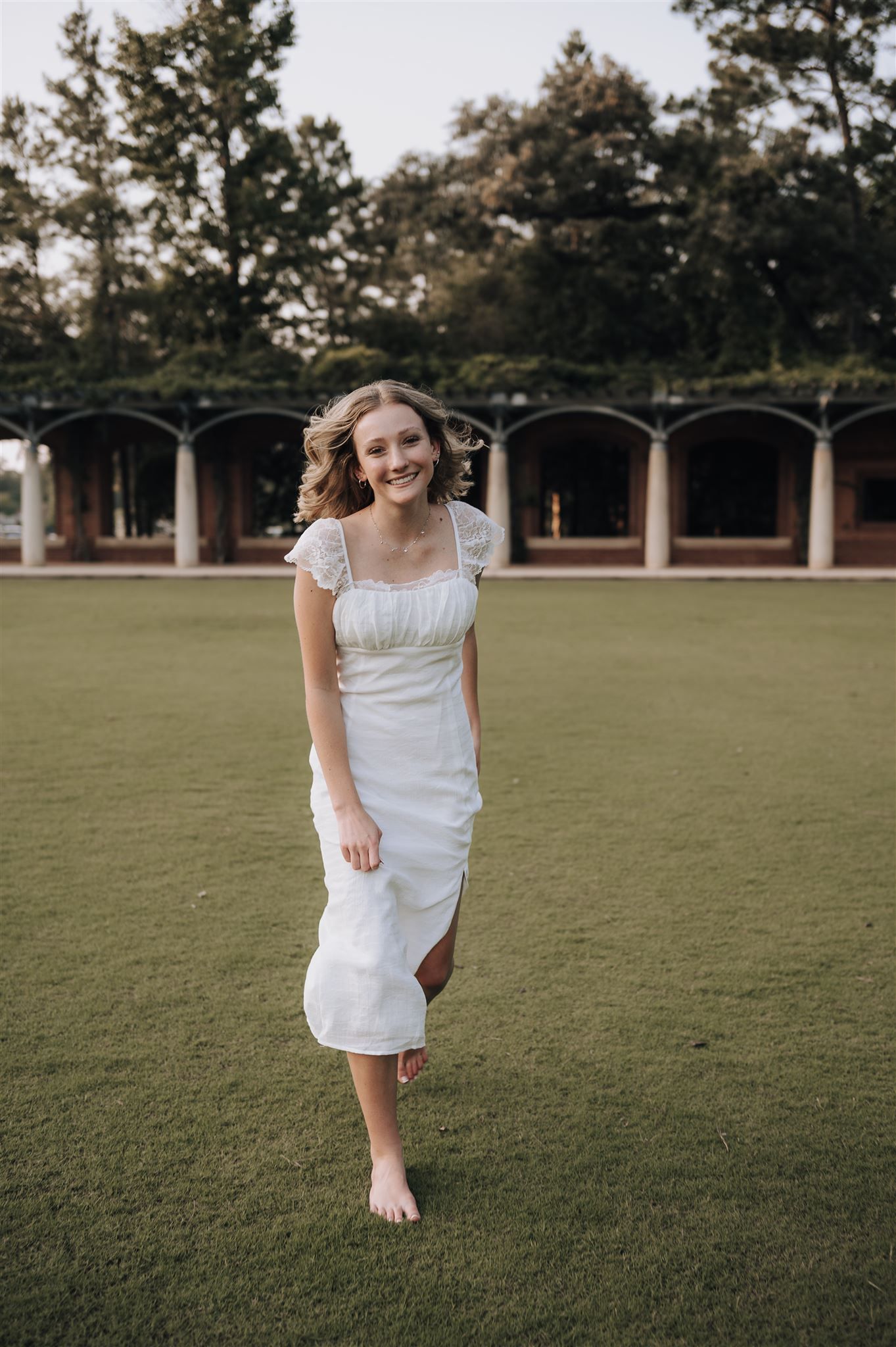 A barefoot person in a white midi dress poses on a green lawn with columned architecture in the background.