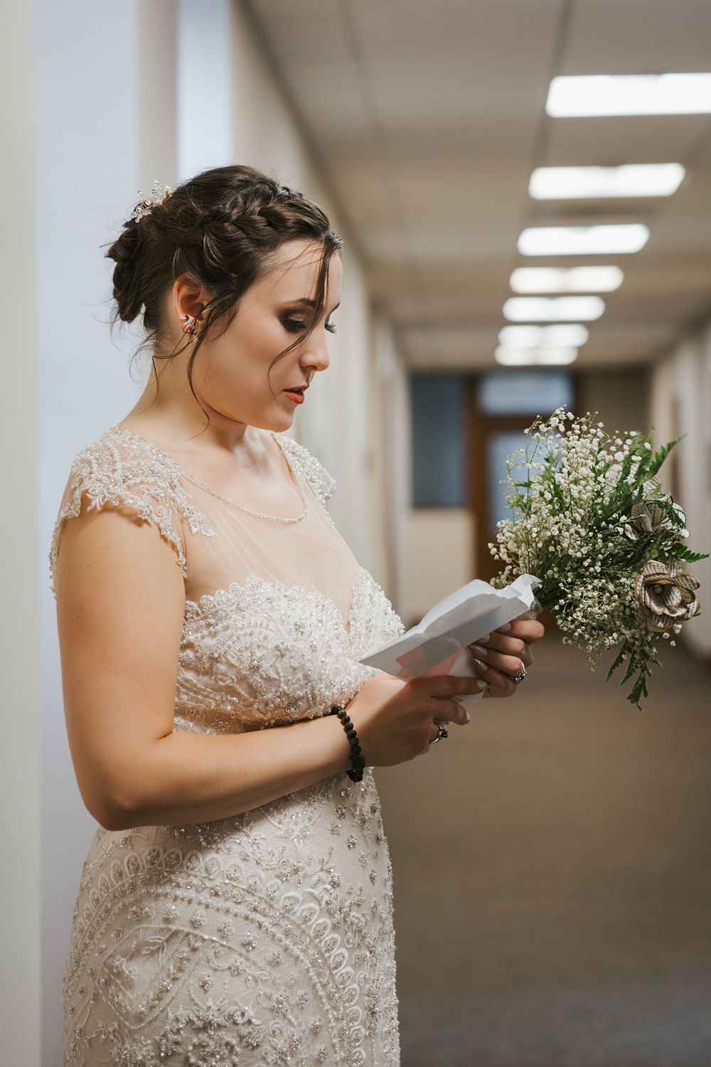 A bride in a lace wedding dress reads a letter while holding a delicate bouquet of baby's breath flowers in a hallway.