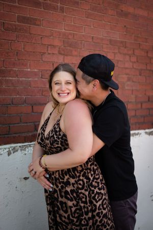 An expecting couple embraces in front of a red brick wall, with the woman wearing an animal print dress.