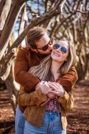 A couple embraces outdoors wearing sunglasses and brown jackets among twisted tree branches in Dallas.