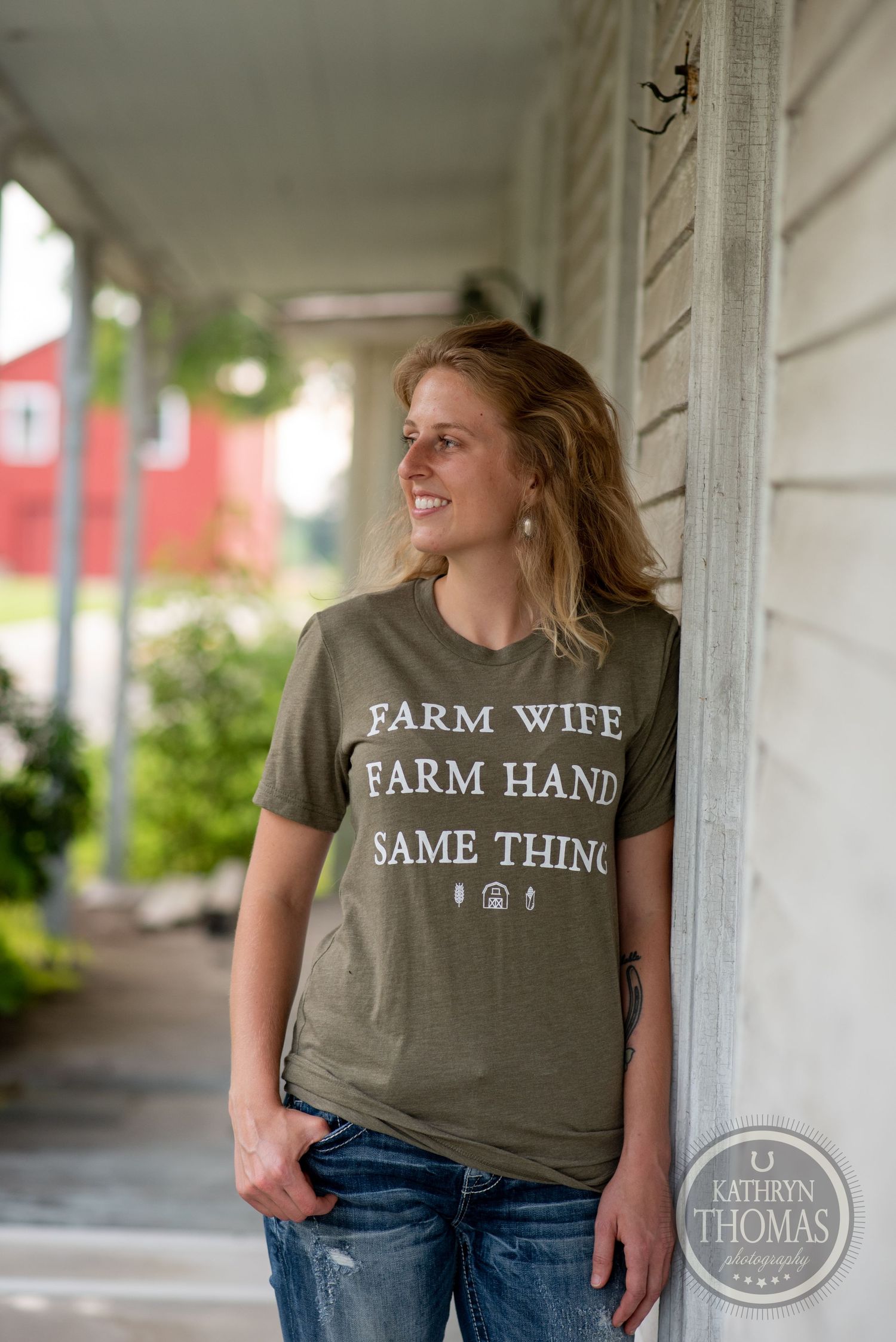 Woman in olive green t-shirt with 'Farm Wife Farm Hand Same Thing' text stands on a farmhouse porch.