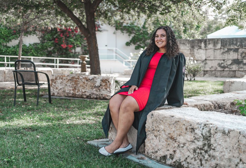 Female graduate in black gown and red dress sitting on stone bench in garden setting.