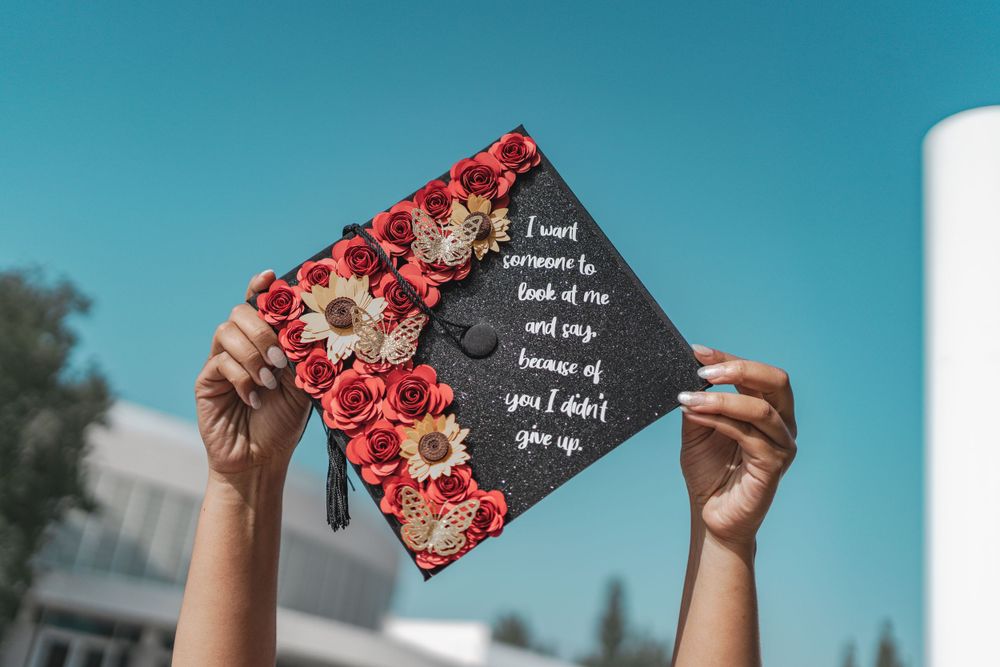 Decorated graduation cap with red roses and sunflowers held up against bright blue sky.