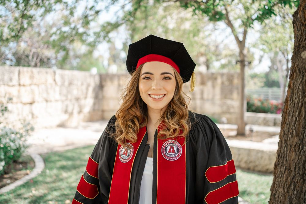 Woman smiles in red and black doctoral graduation cap and gown among trees outdoors.