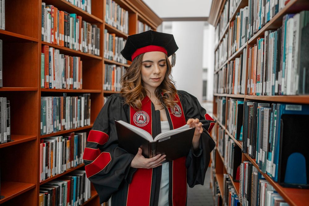 Female doctoral graduate reads book in library aisle wearing red and black graduation regalia.