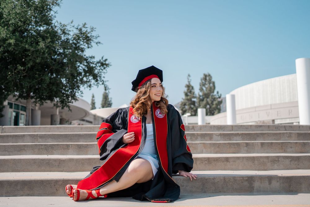 Female doctoral graduate in red and black regalia sits relaxed on outdoor campus steps.
