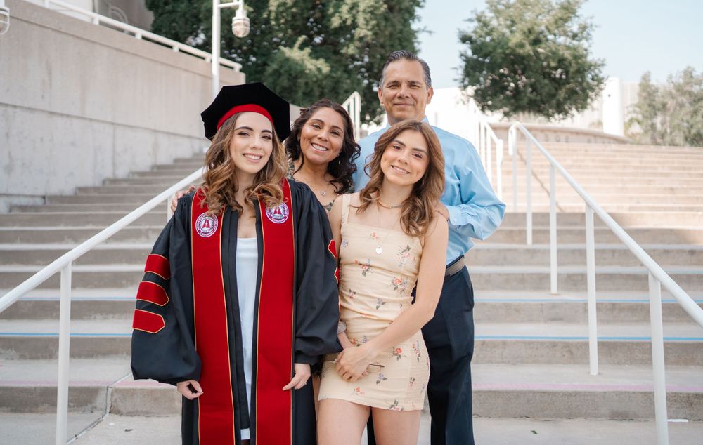 Doctoral graduate in red regalia poses with family on outdoor steps after graduation ceremony.