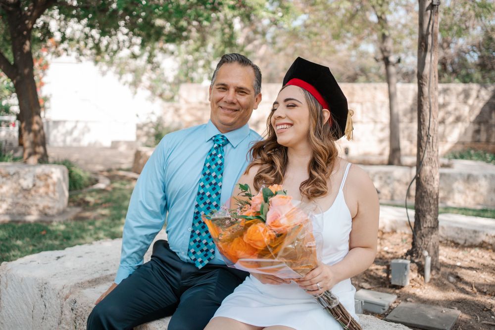 Young female graduate in cap holds flowers bouquet sitting with older man in blue tie outdoors.