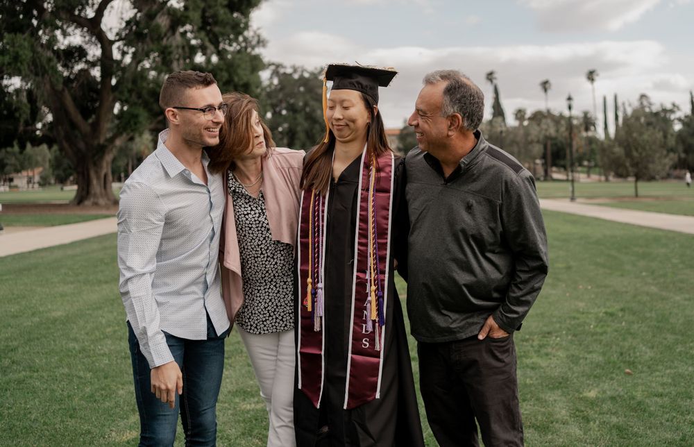 Graduating student in cap and stole celebrates with smiling family group on green campus lawn.