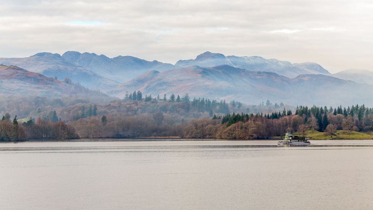 Scenic mountain landscape with misty hills and a lake in the foreground under cloudy skies.