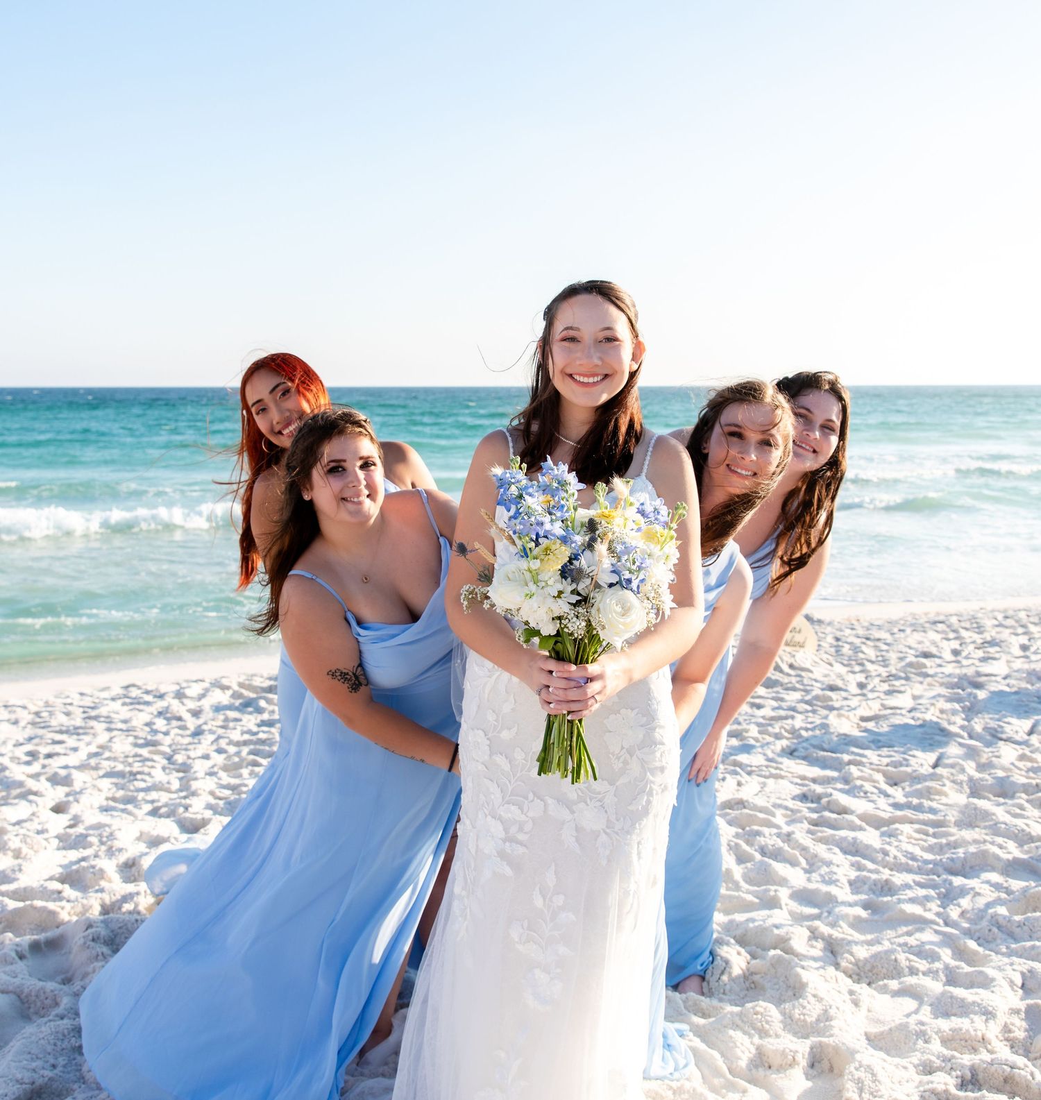 A bride and her bridesmaids pose on a beach, holding bouquets.