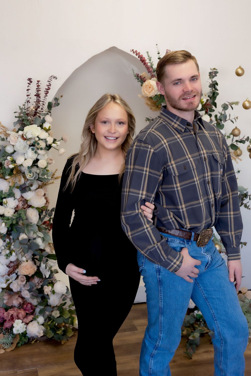 Maternity photo with floral arch featuring couple in black dress and plaid shirt.
