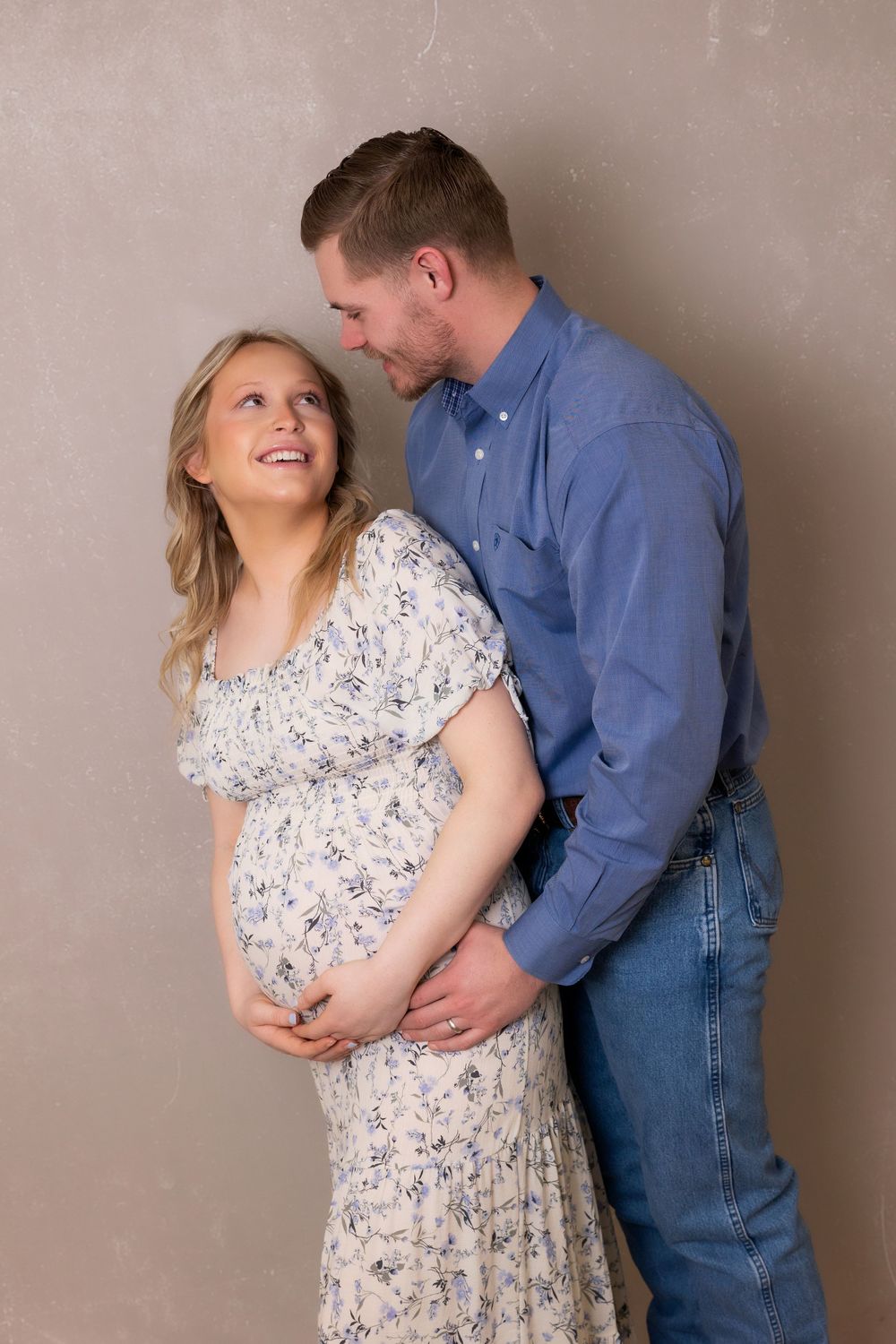 A joyful maternity portrait showing a couple sharing a loving moment against a neutral wall.