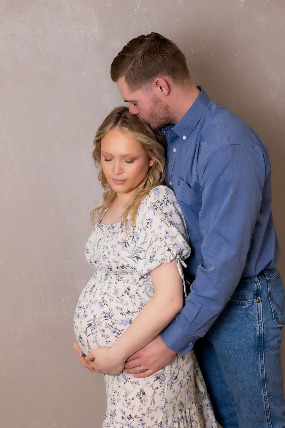 An intimate maternity photo captures a tender moment between a couple against a grey backdrop.