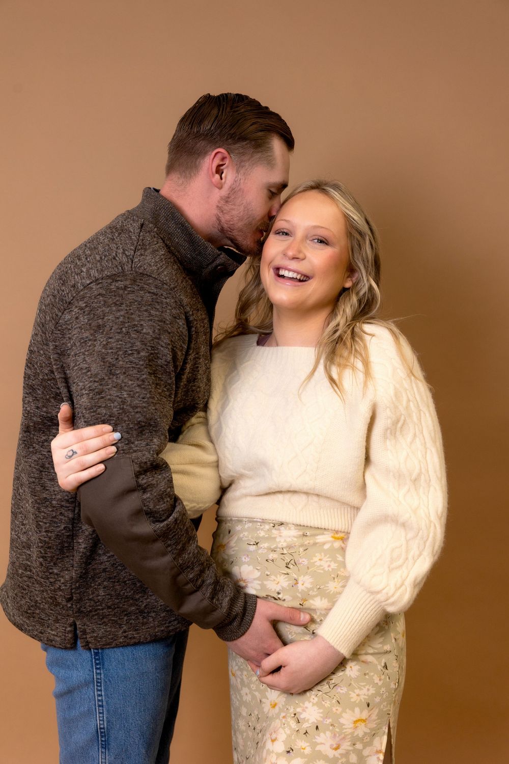 An affectionate couple shares a laugh during their maternity photoshoot against a brown background.