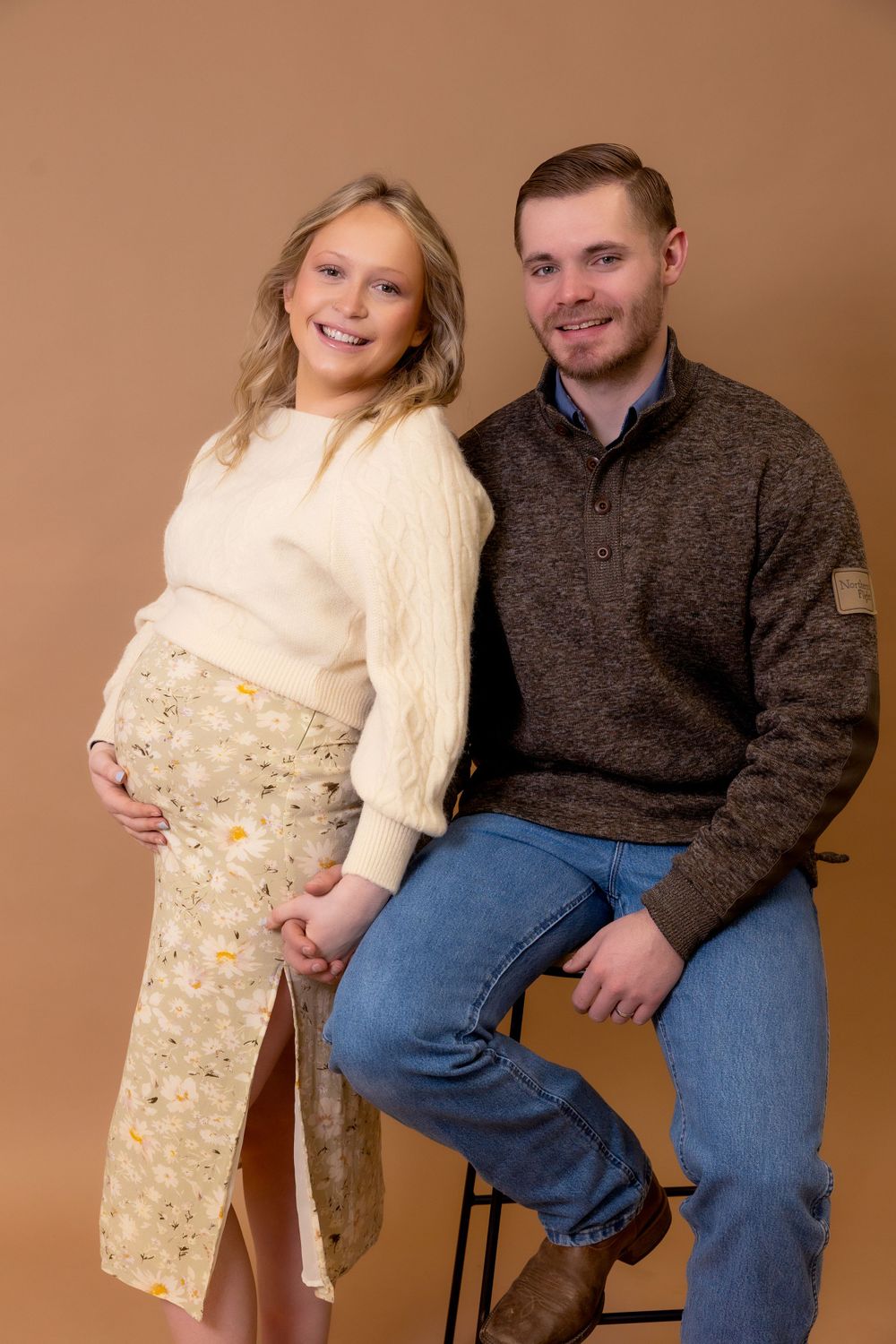 A maternity photoshoot featuring a couple in cozy sweaters against a warm brown backdrop.