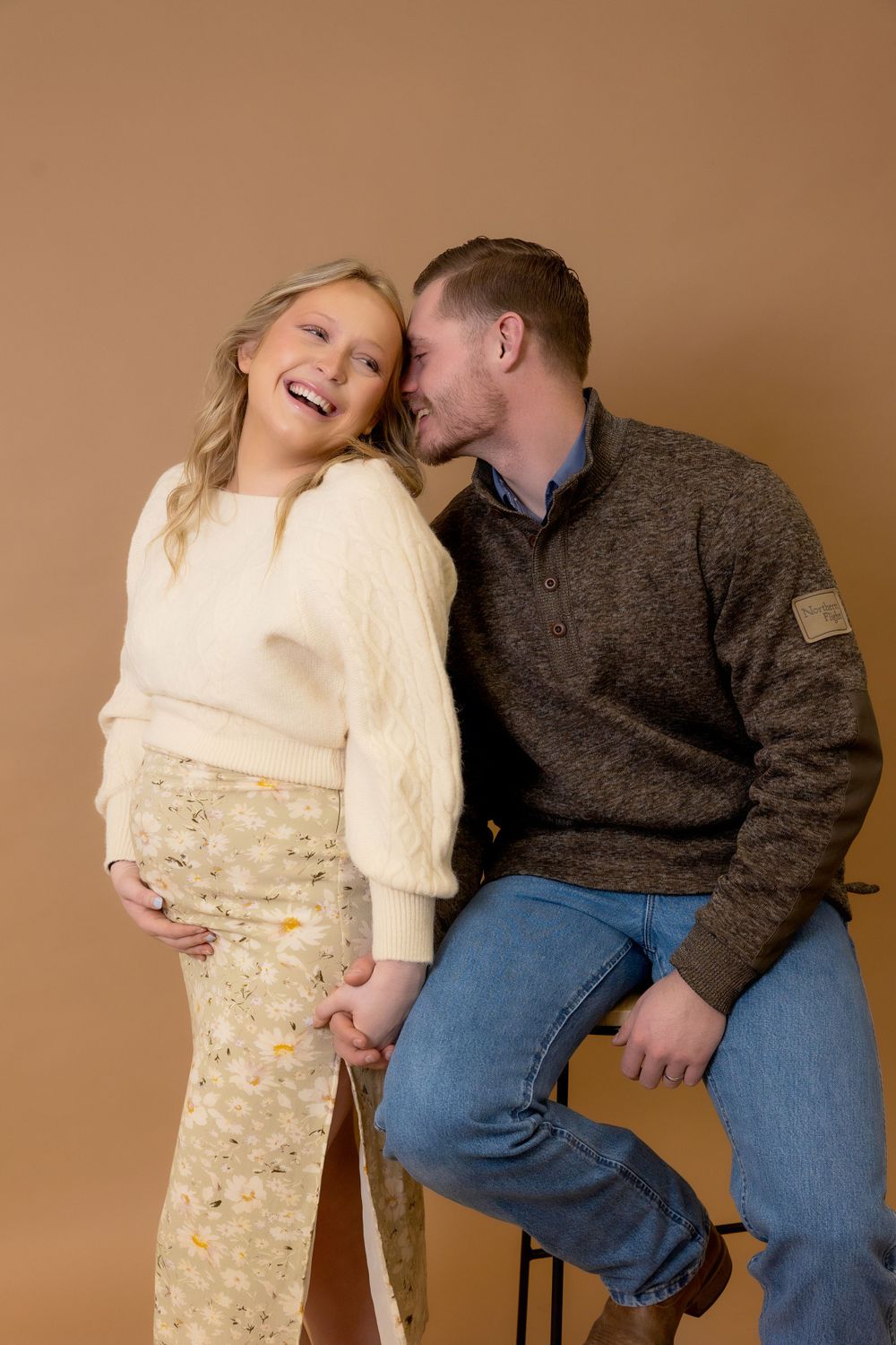 A joyful maternity photo session with a couple in casual winter attire against a neutral backdrop.