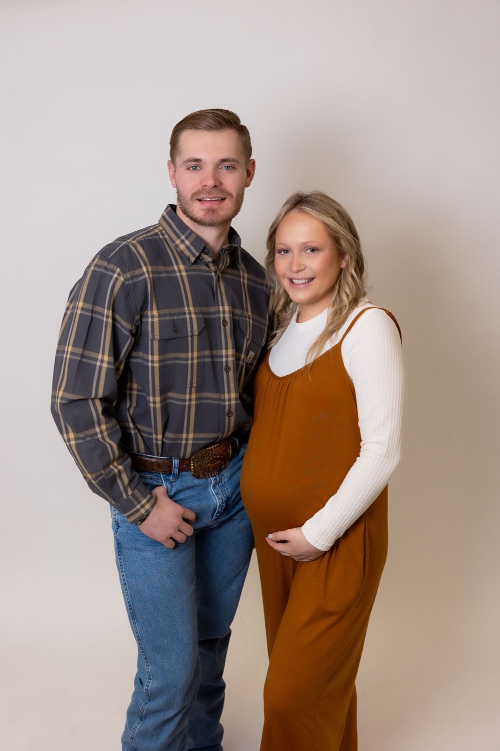 Couple poses for maternity photo in casual attire against neutral backdrop.