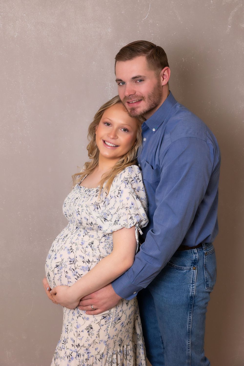 A couple poses for a maternity photo in classic attire against a textured grey wall.