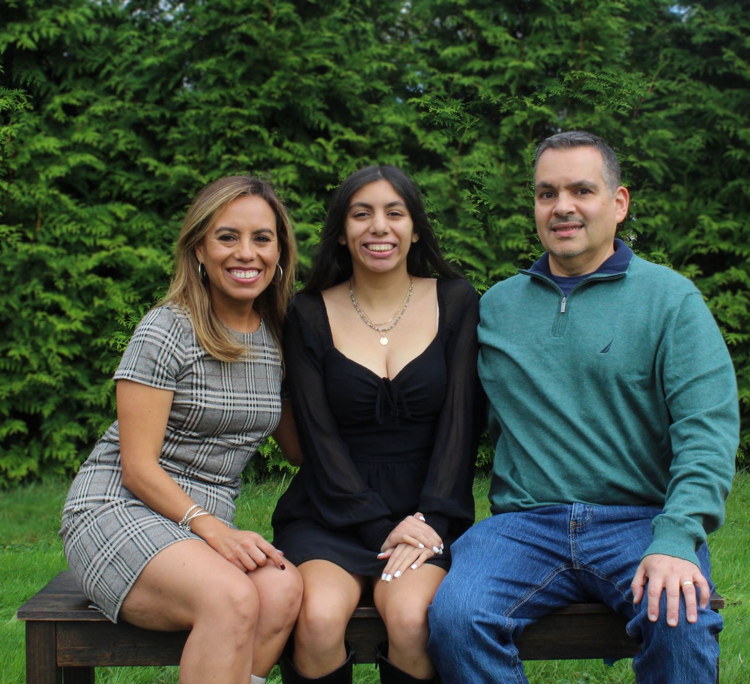Three people sitting together on wooden bench with green hedge background.