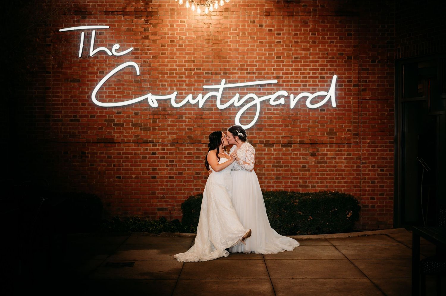 Two brides sharing first dance kiss under neon Courtyard sign at wedding reception.