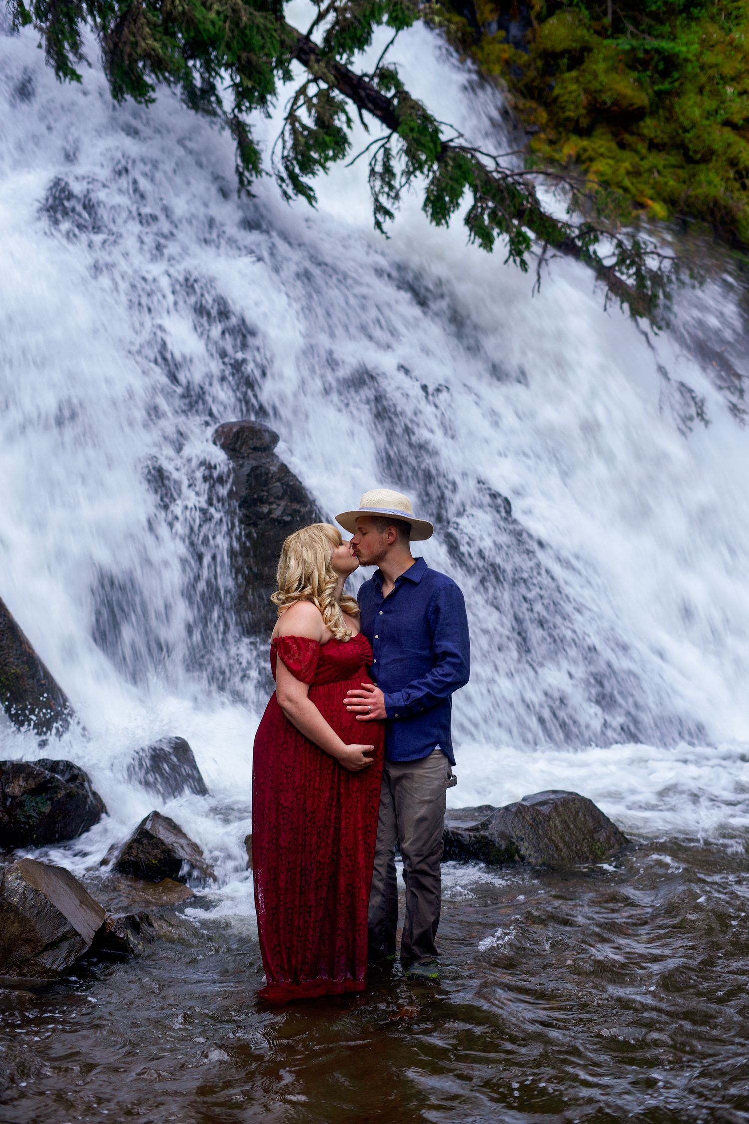 Couple stands in waterfall stream during romantic maternity photoshoot.