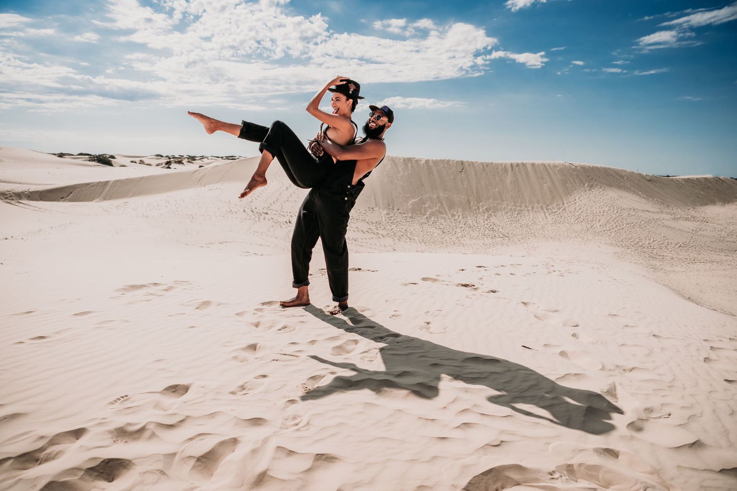 A playful couple enjoying a sunny day at white sand dunes against a bright blue desert sky with puffy clouds.
