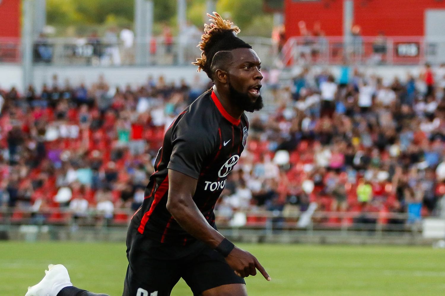 Soccer player celebrates emotionally during a match wearing a black uniform in front of stadium crowd.