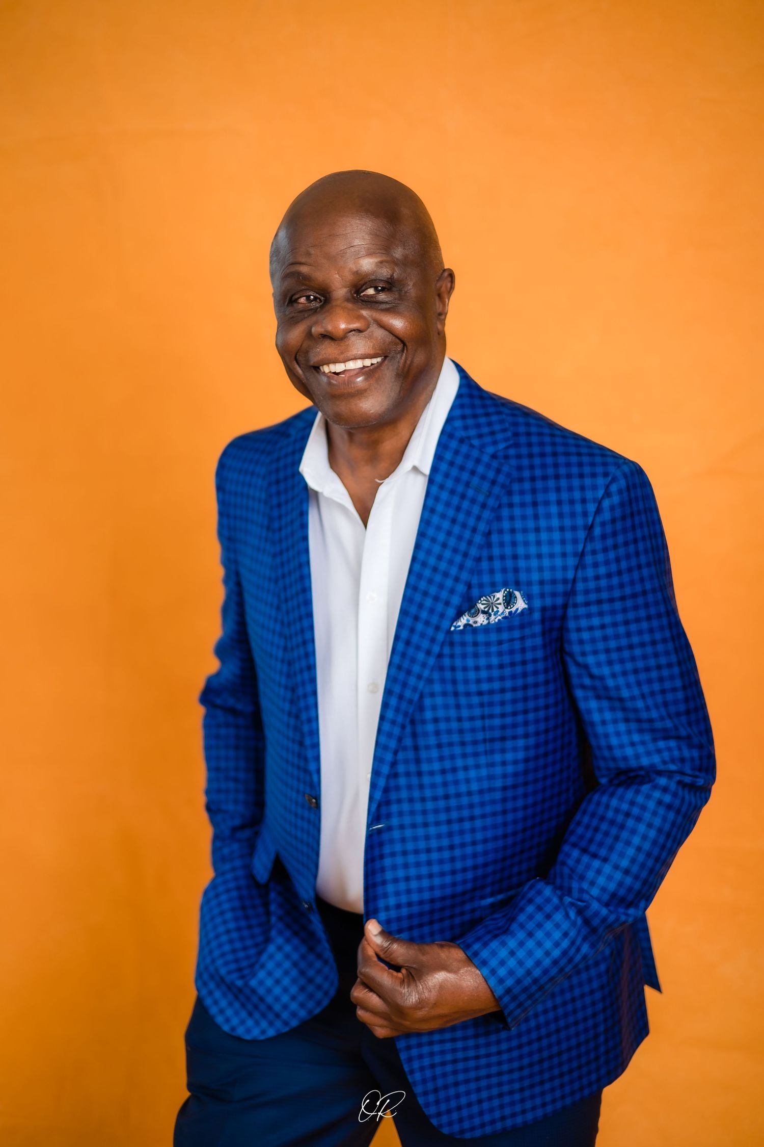 A distinguished gentleman in a checkered blue blazer and white shirt poses against an orange background.