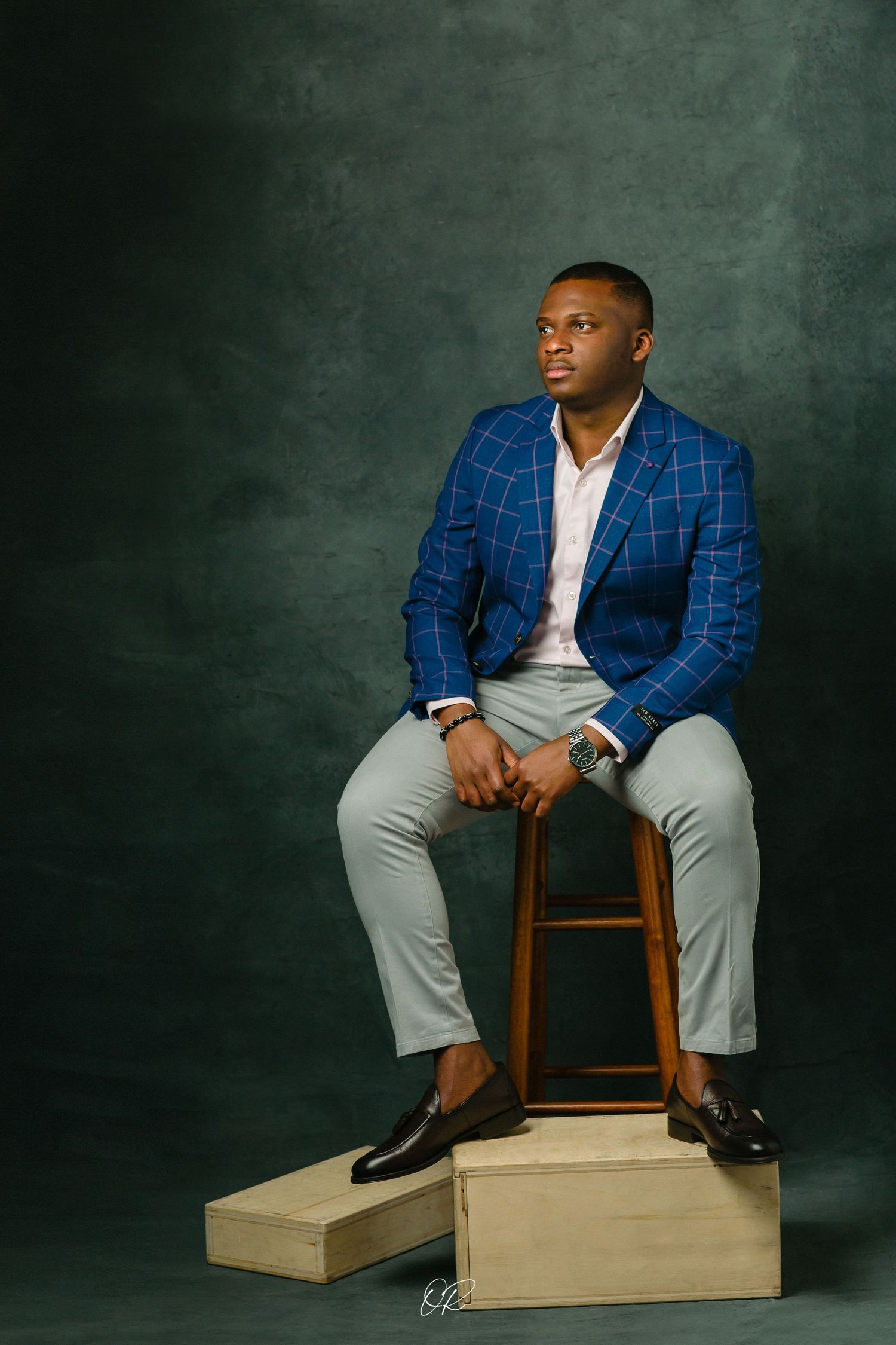 Portrait of person in blue checkered blazer sitting on wooden stool against dark background.