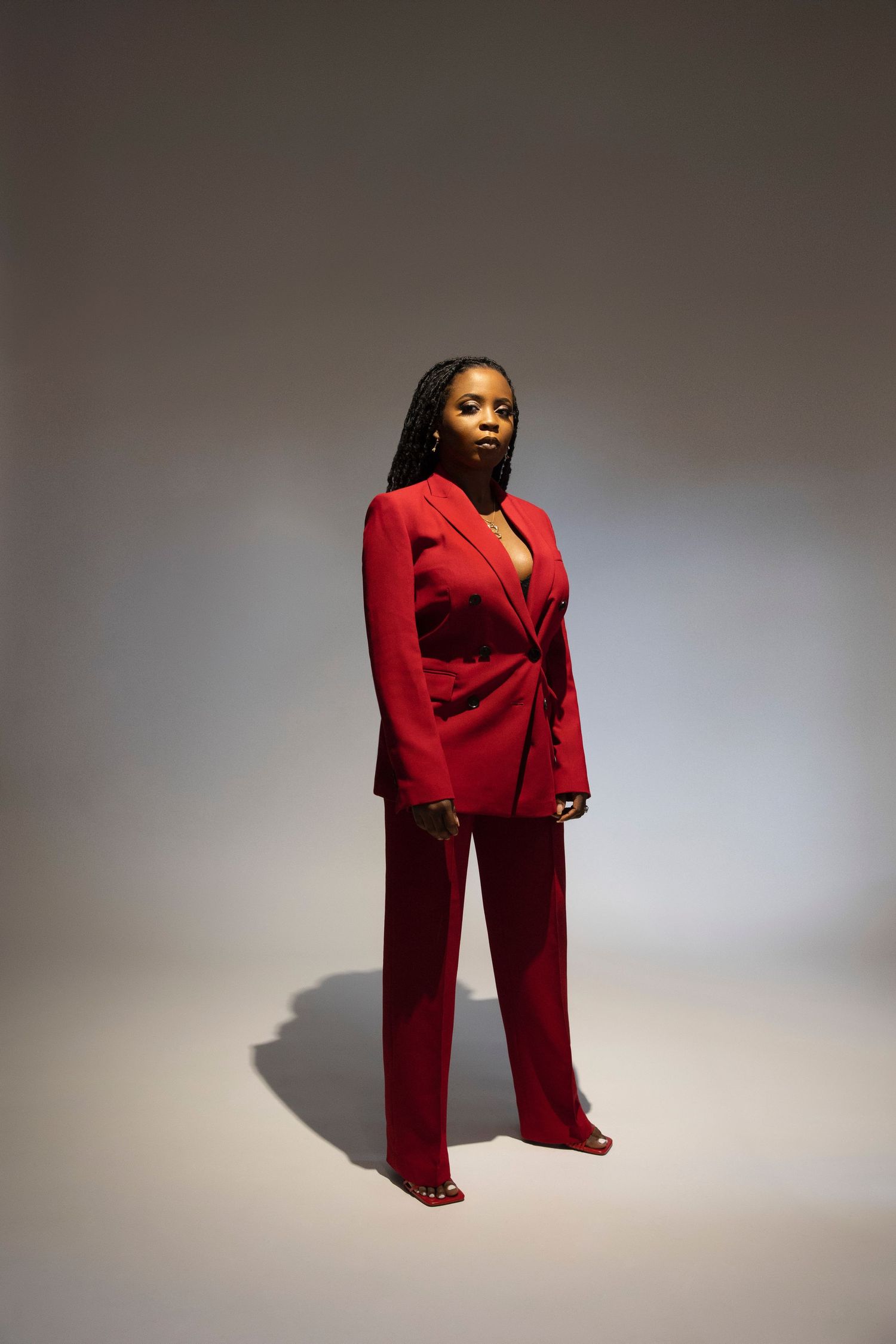 A fashion model stands powerfully in a red pantsuit against a dramatic studio backdrop.