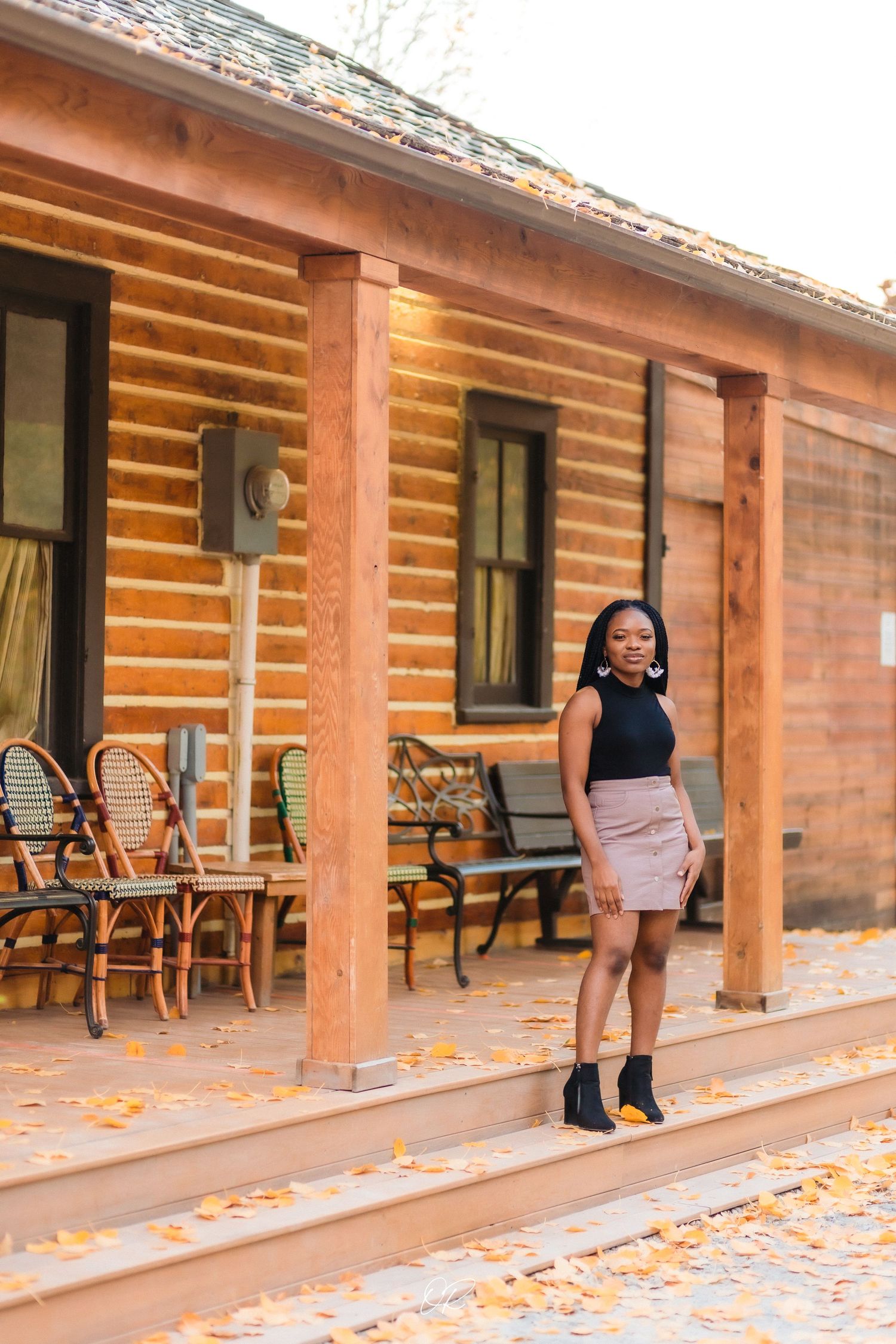 Person in black top poses on wooden porch with autumn leaves scattered around.