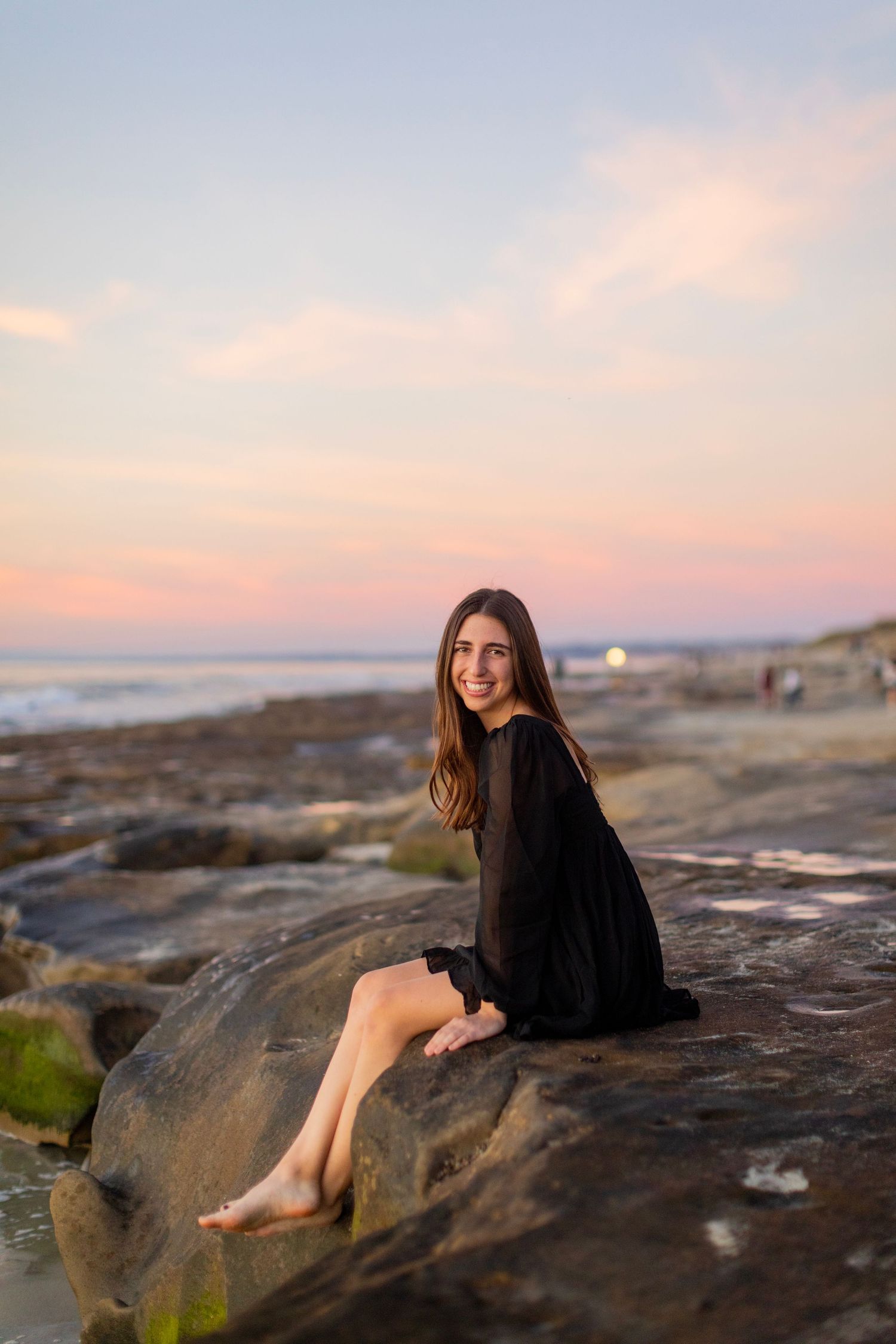 A high school senior in a black dress sits on coastal rocks during sunset at the beach with pink and orange sky in the background.