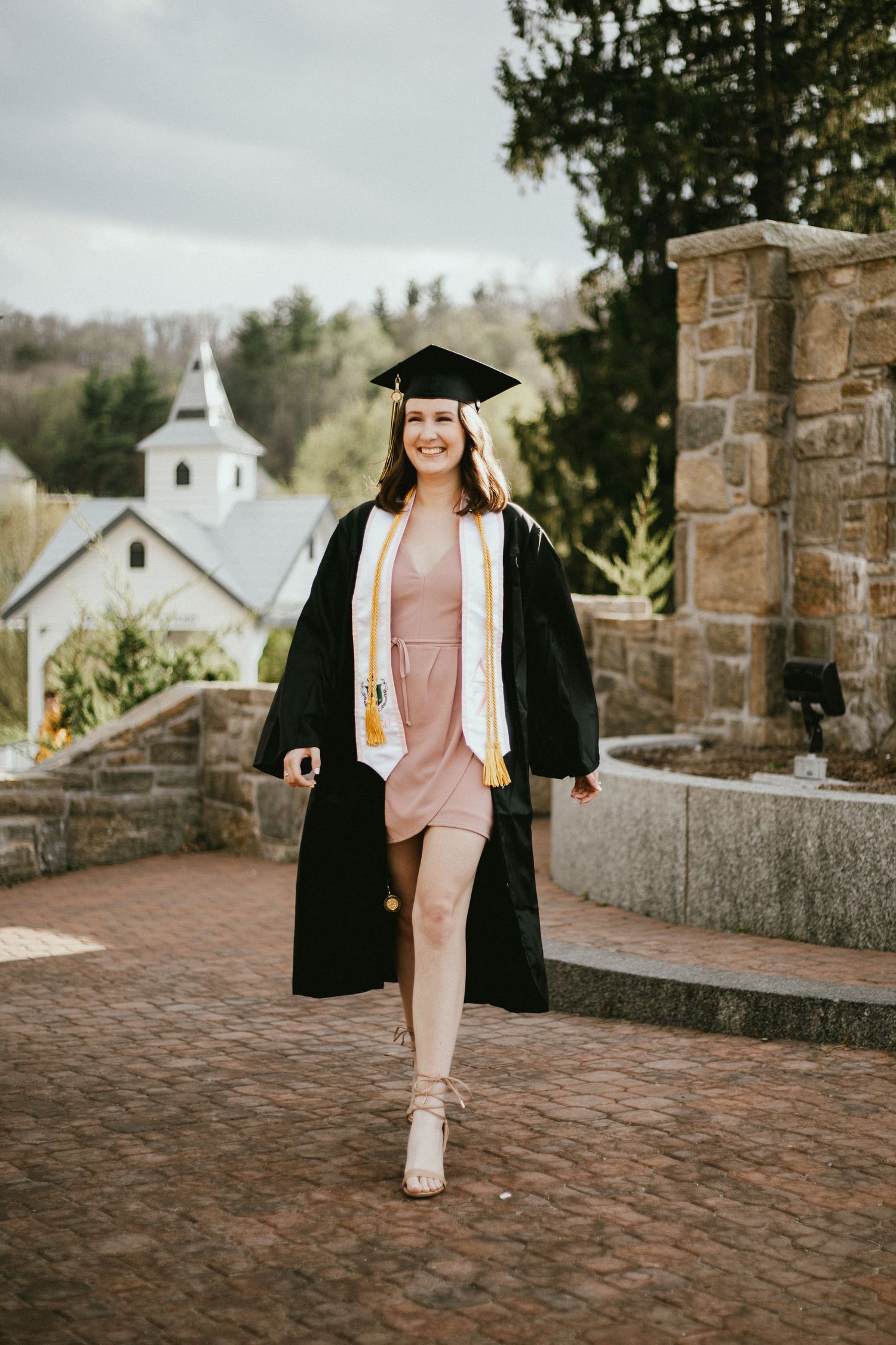 Graduate in pink dress and black cap and gown poses in front of stone church building.