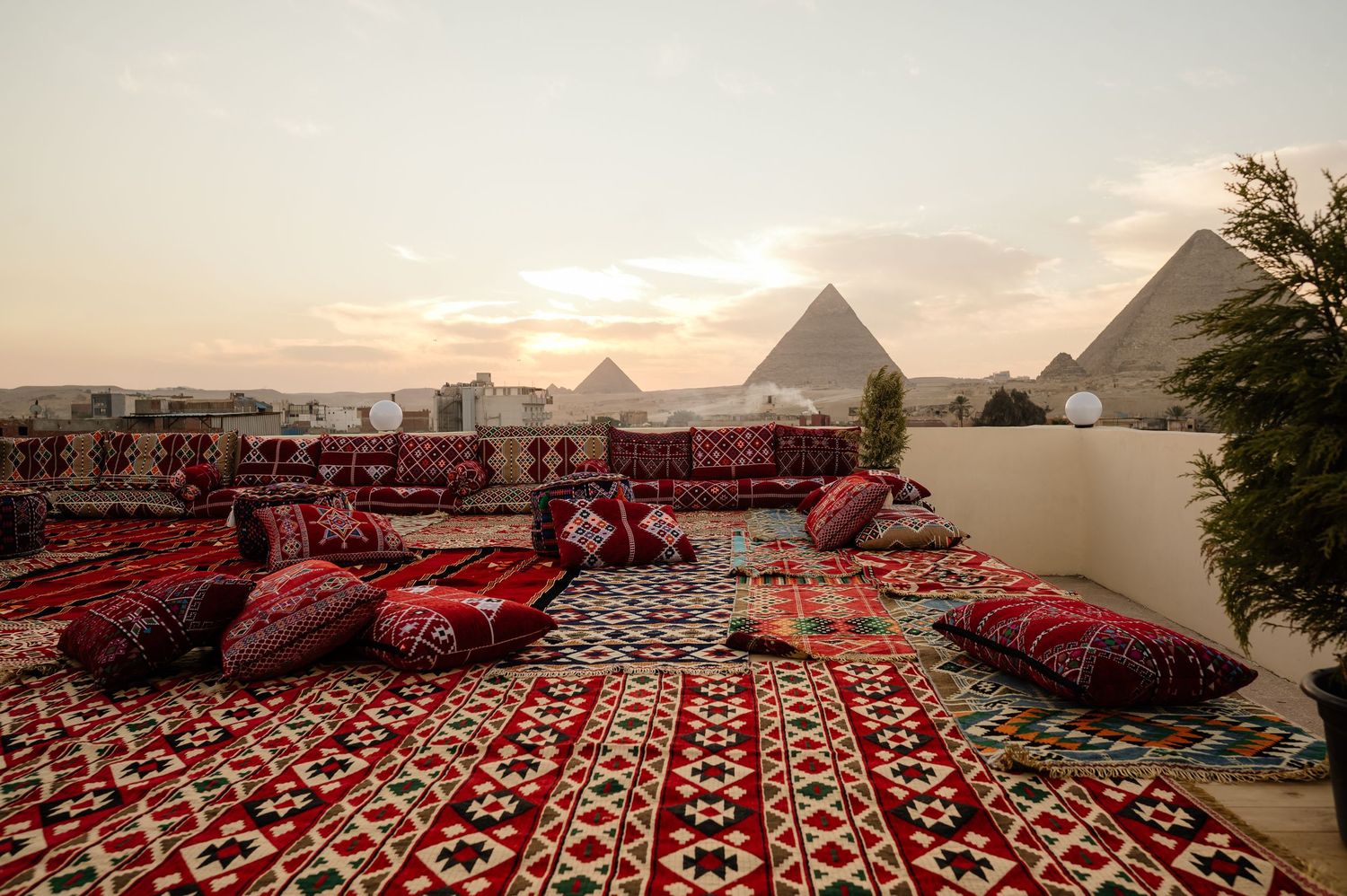 Rooftop terrace with red patterned carpets and cushions overlooking the pyramids at sunset.